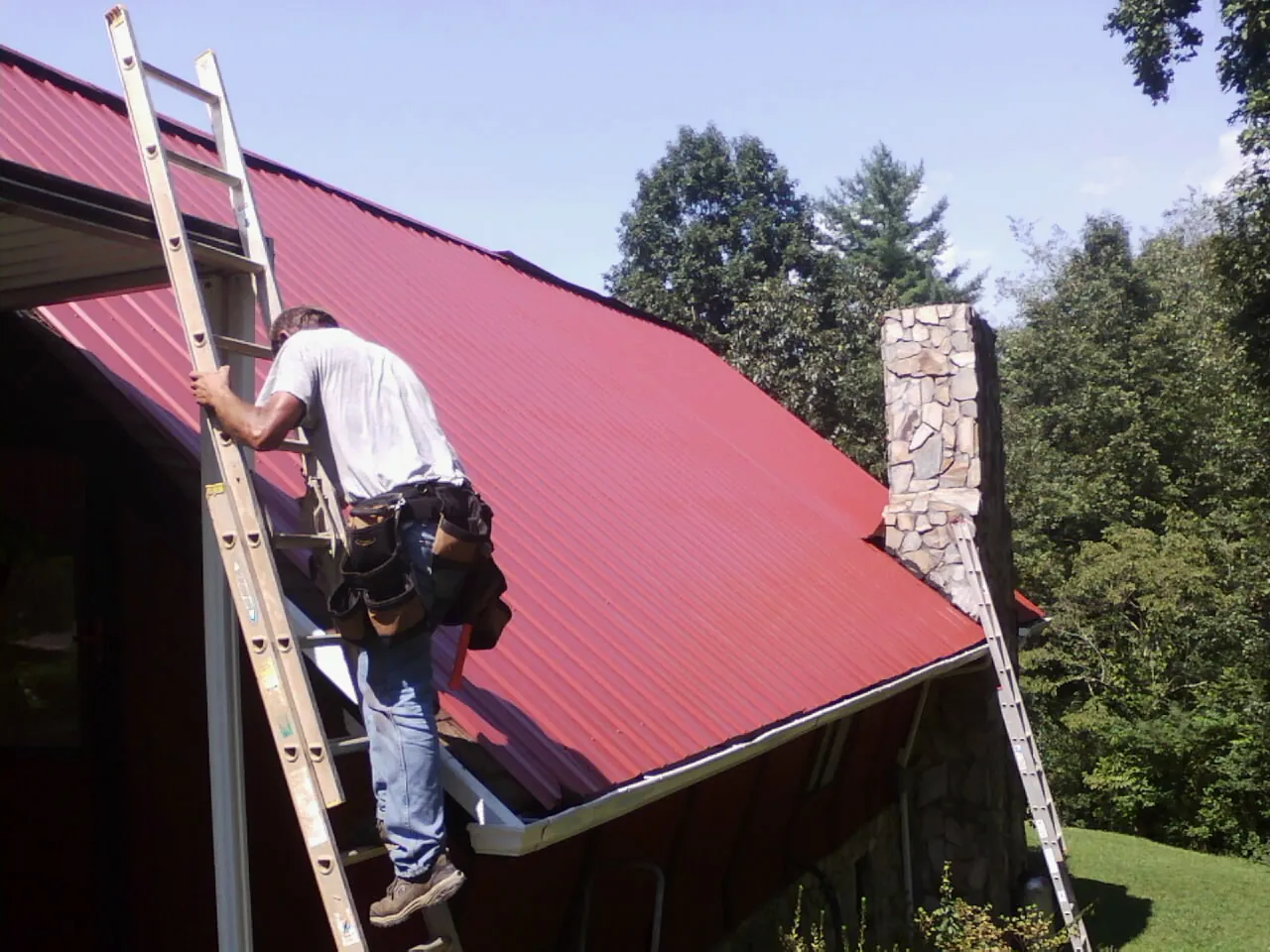 Roofer on a ladder beside a red metal roof and stone chimney — Soffit & Fascia Repair by Roofing Brothers in Danbury